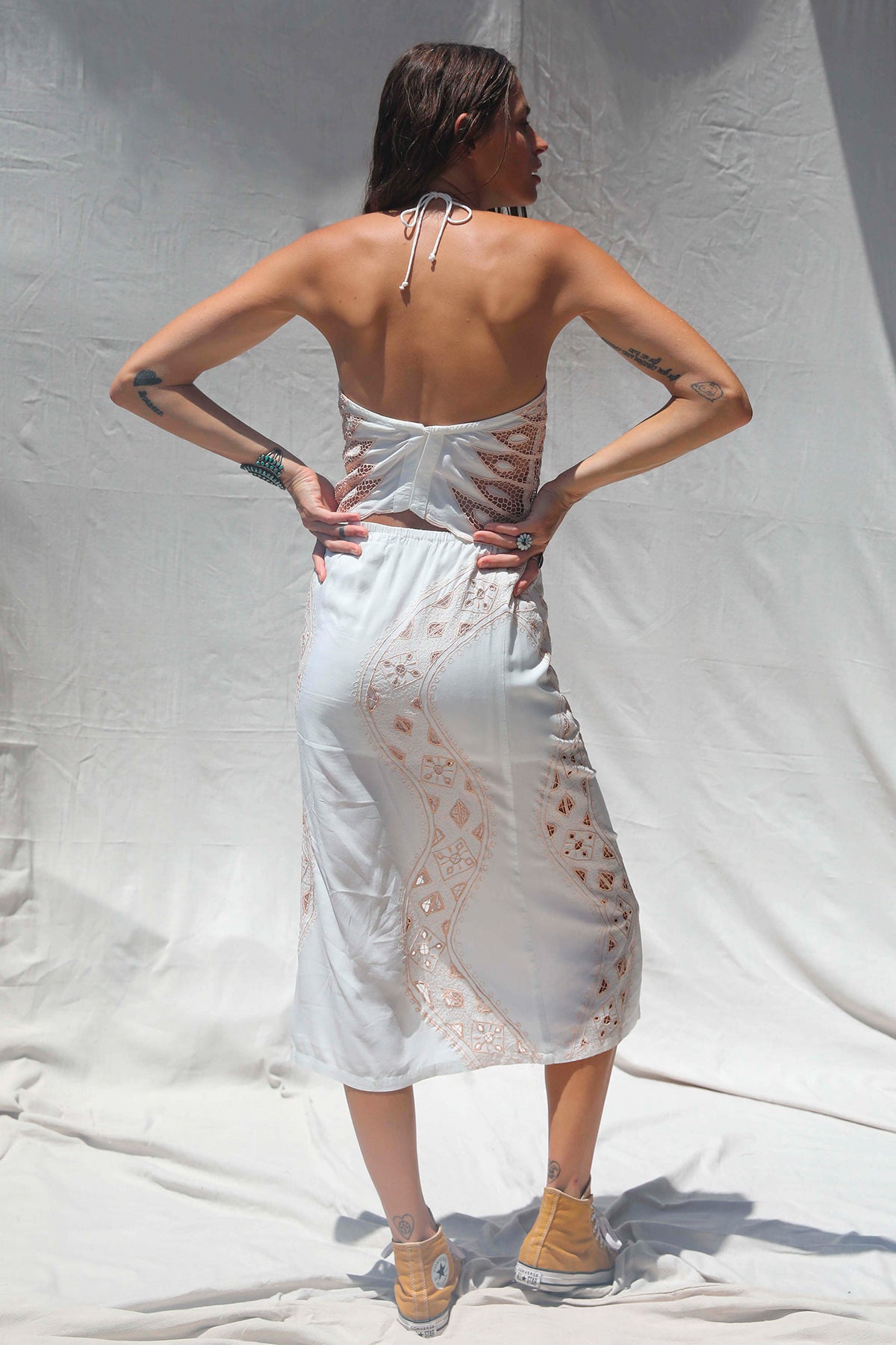 Modelo de pie de espaldas vistiendo un vestido blanco con bordados y detalles transparentes, posando sobre una tela blanca