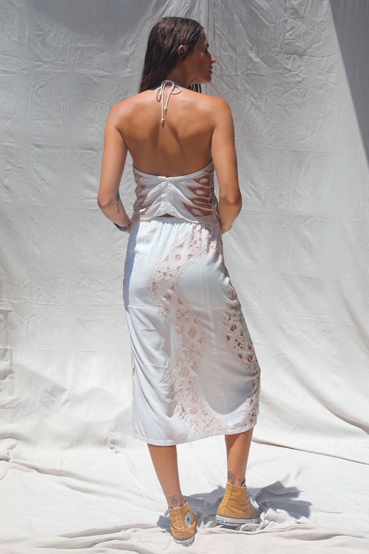 Modelo de espaldas con vestido blanco bordado con detalles calados, corte midi y tirantes finos estilo halter en exterior al aire libre