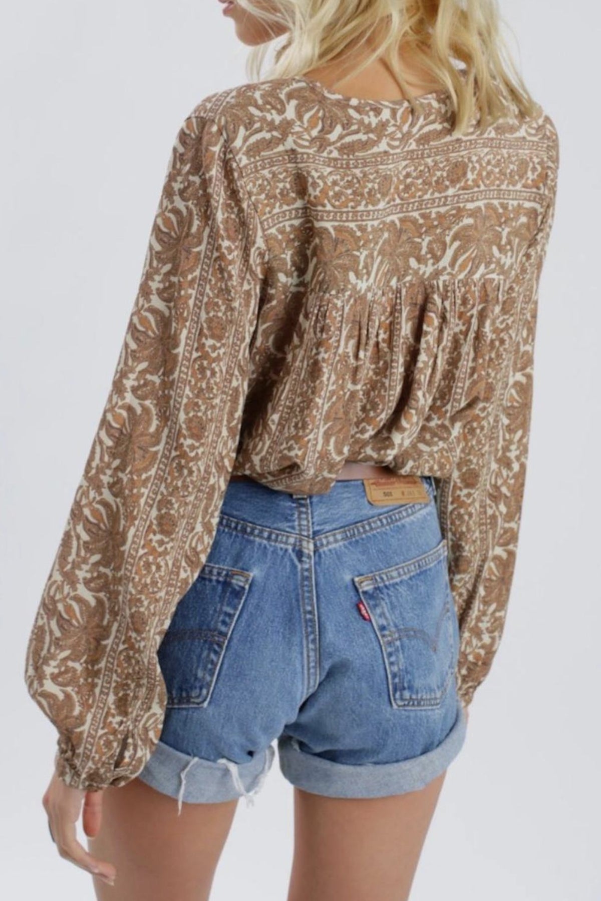 Modelo posando de espaldas con blusa marrón Sunset Meadow, mostrando el cierre de lazo y caída fluida sobre fondo blanco limpio