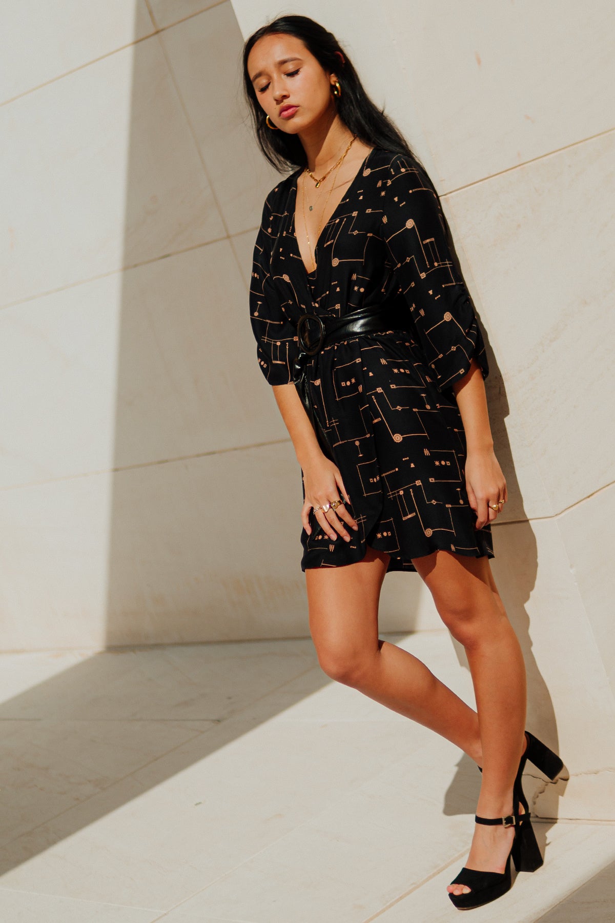 Modelo apoyada en pared con vestido Eskell Esme negro y estampado dorado, destacando el corte envolvente y silueta estilizada sobre fondo neutro