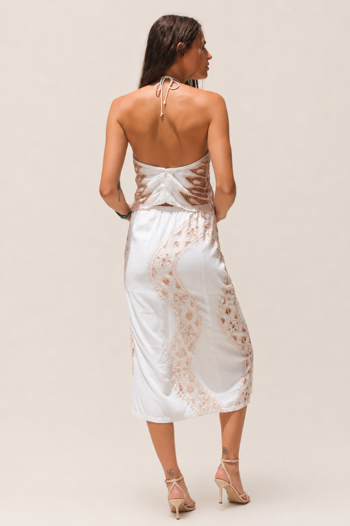Modelo de espaldas con vestido blanco bordado con detalles calados, corte midi y tirantes finos estilo halter en exterior al aire libre